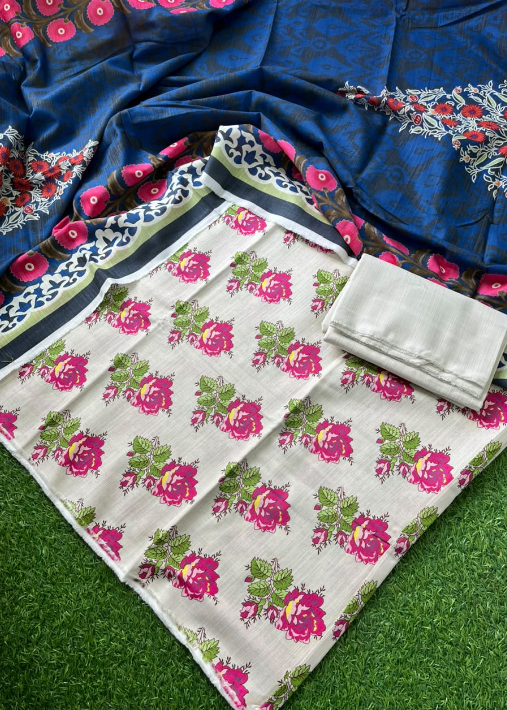A folded floral fabric set laid out on green artificial grass, featuring a cream-colored base with pink and green floral print, a matching blue patterned dupatta with red and white floral borders, and a folded light gray fabric piece.
