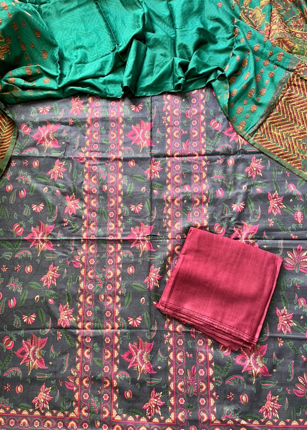 A folded and draped salwar suit set laid flat, featuring a dark gray printed fabric with pink and green floral motifs, a bright green dupatta with gold patterns, and a folded pink fabric piece.