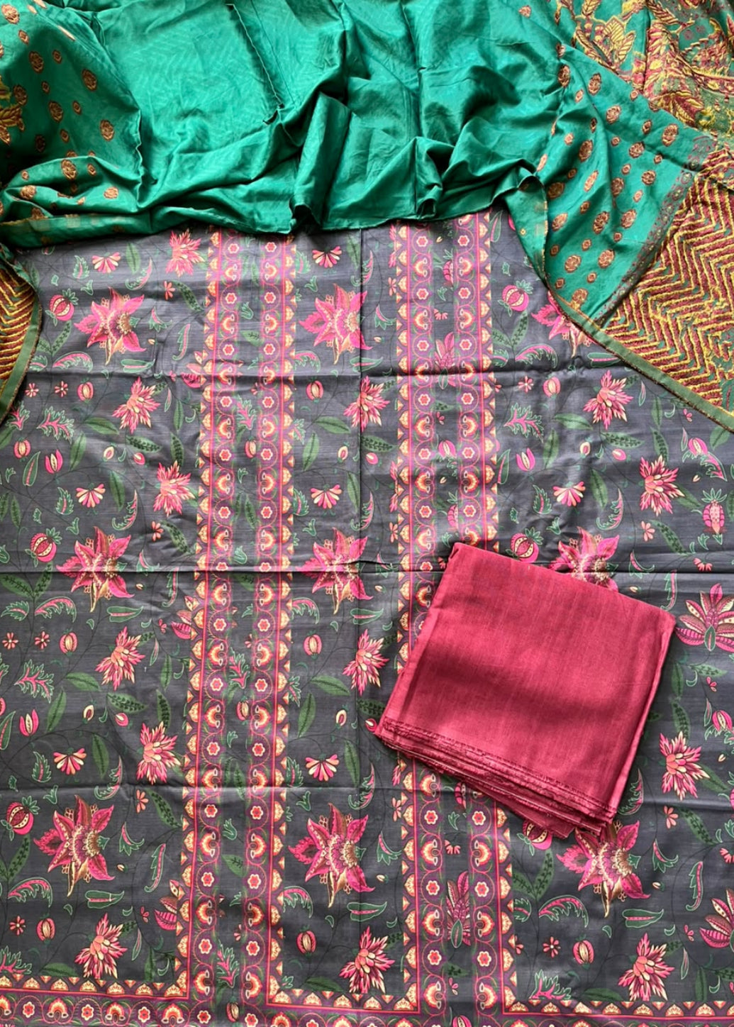 A folded and draped salwar suit set laid flat, featuring a dark gray printed fabric with pink and green floral motifs, a bright green dupatta with gold patterns, and a folded pink fabric piece.