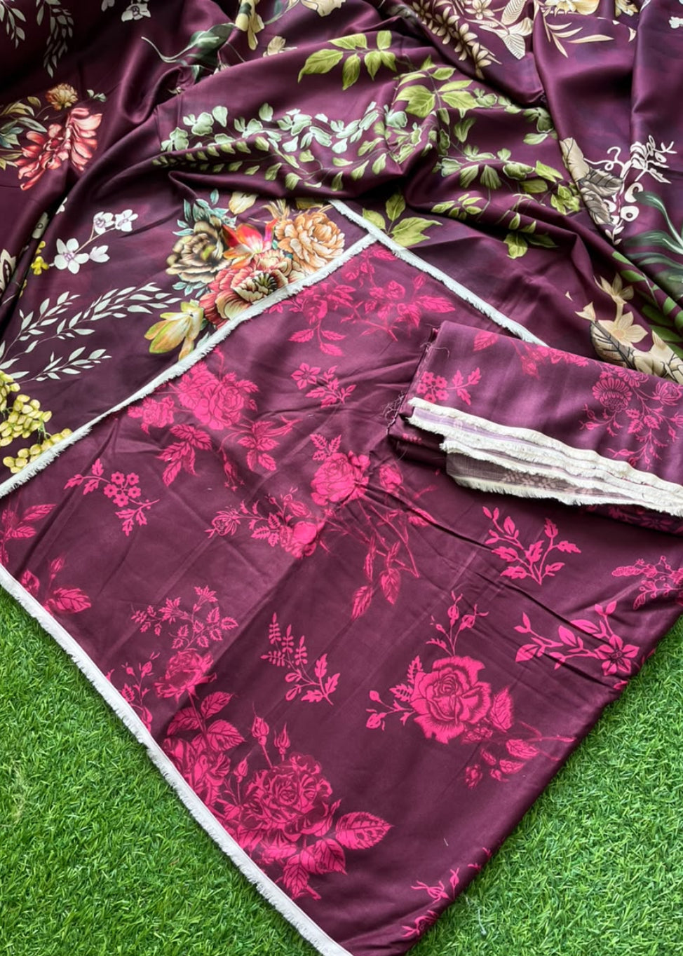 A draped piece of deep magenta-purple fabric with a pink floral print and white border trim, laid out on green artificial grass. A folded matching fabric piece with a lace trim is placed on top.