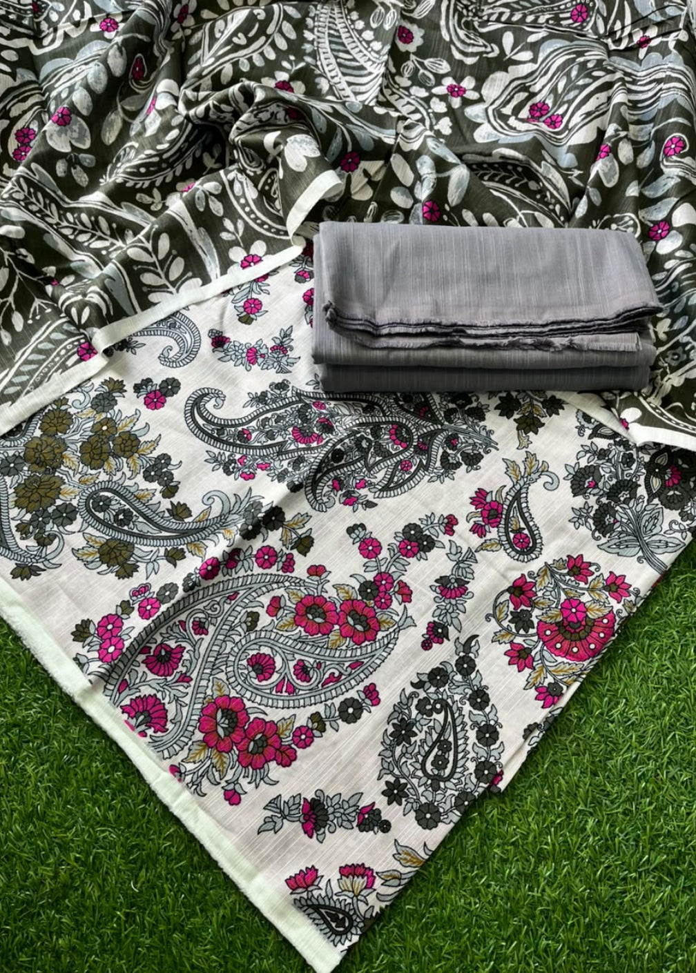 A folded and unfolded piece of printed fabric laid on artificial green grass. The fabric has a white base with gray and black paisley and floral motifs, plus bright pink floral accents, with a folded gray fabric piece placed on top.