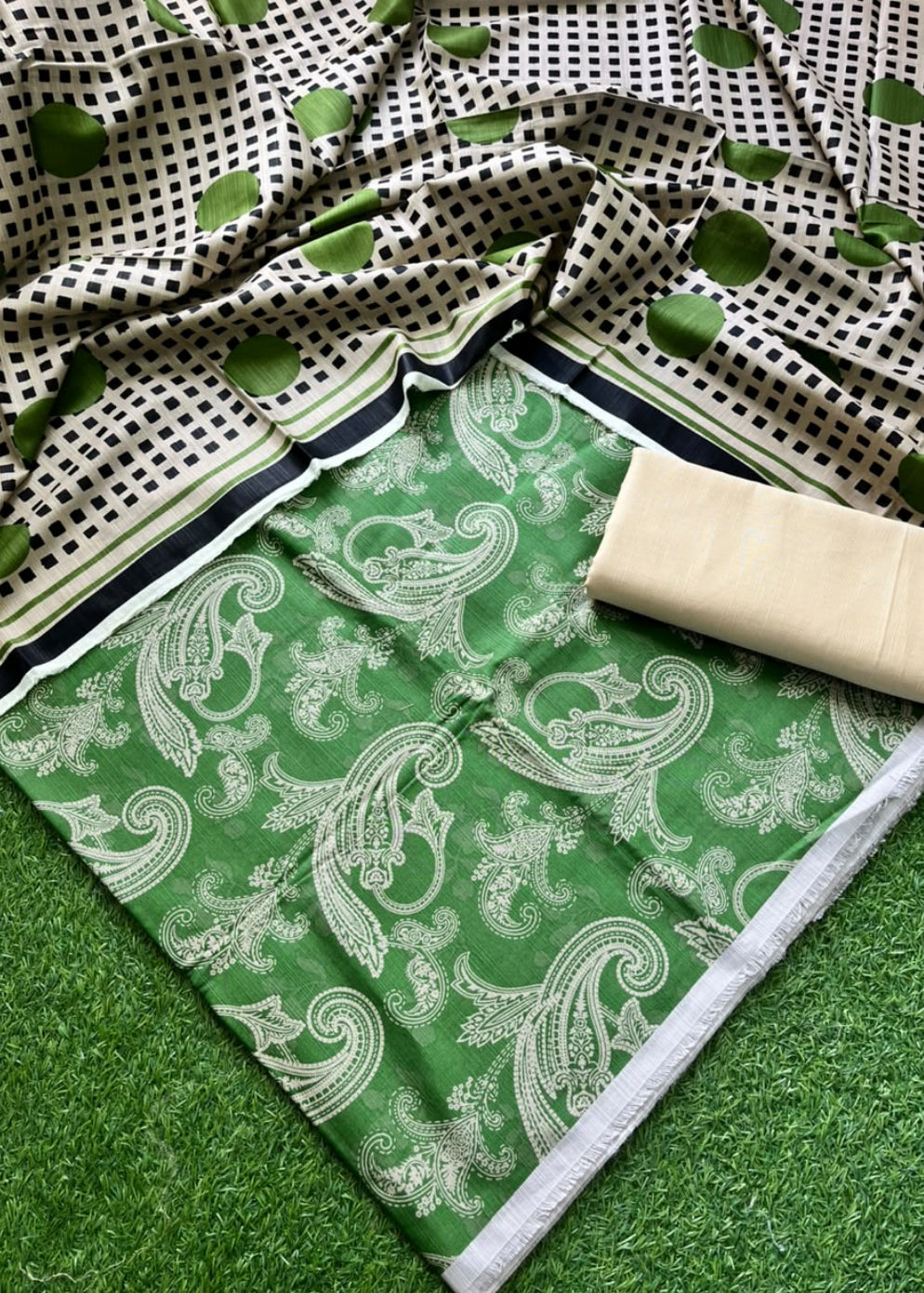 A green and white printed fabric set laid out on artificial grass, featuring a paisley patterned main section, a black-and-white dotted dupatta, and a folded beige fabric piece.