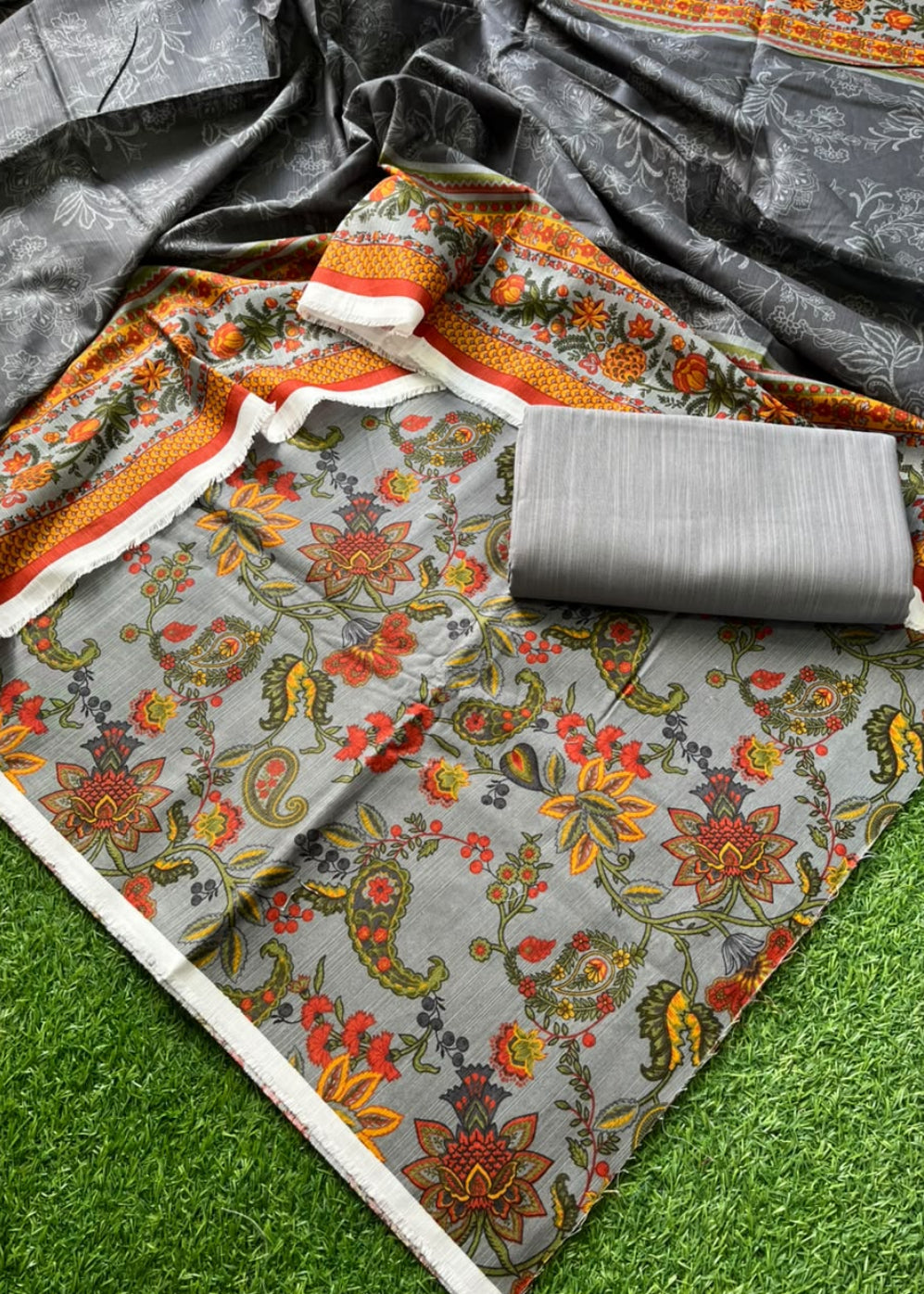 A folded and draped saree fabric set on artificial green grass, featuring a gray base with a colorful floral and paisley print in orange, red, yellow, and green, along with a matching gray fabric roll.