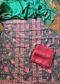 A folded and draped salwar suit set laid flat, featuring a dark gray printed fabric with pink and green floral motifs, a bright green dupatta with gold patterns, and a folded pink fabric piece.