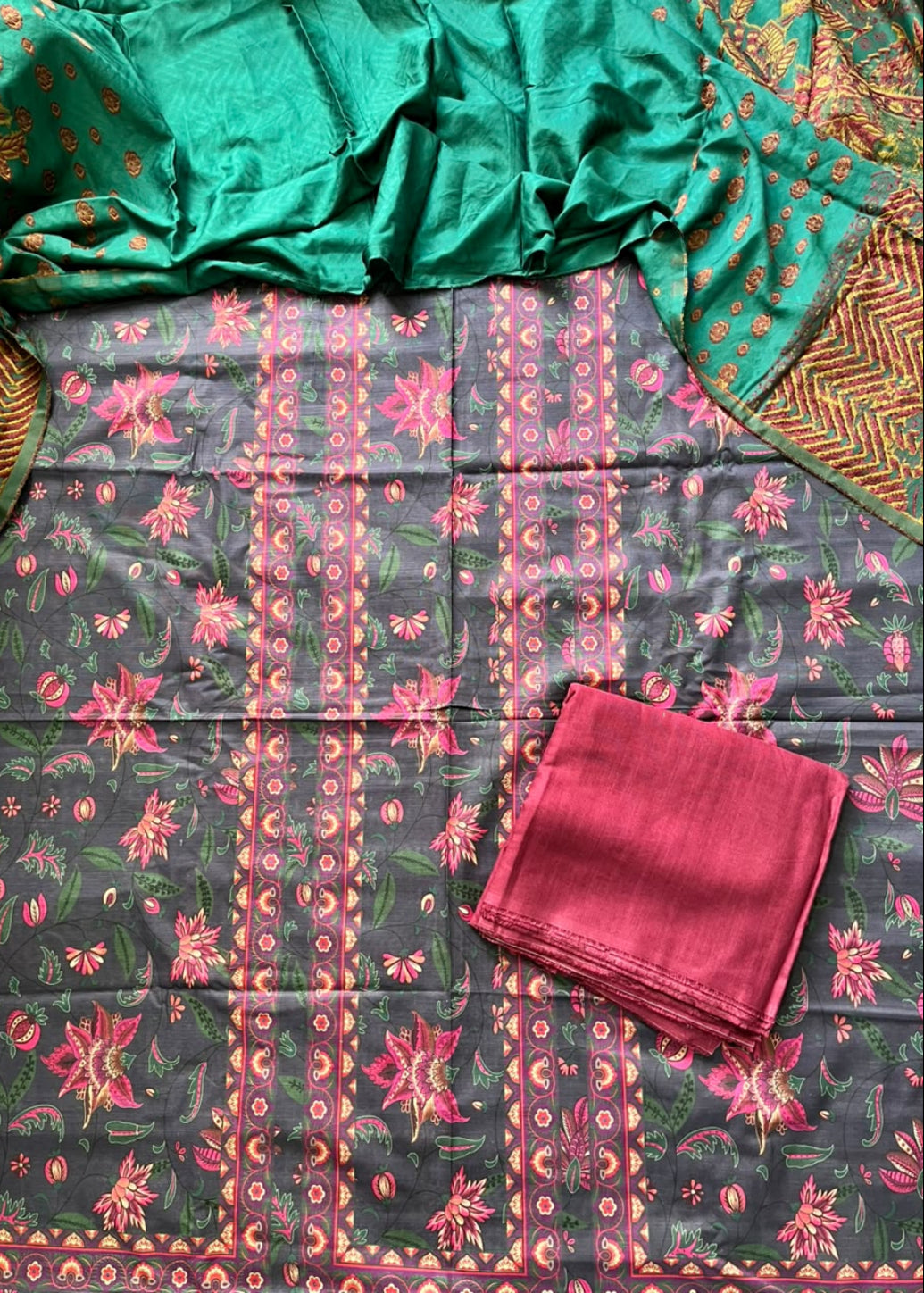 A folded and draped salwar suit set laid flat, featuring a dark gray printed fabric with pink and green floral motifs, a bright green dupatta with gold patterns, and a folded pink fabric piece.