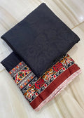A folded dark charcoal gray fabric piece with a subtle jacquard pattern, layered over a matching dark fabric and a red, black, and multicolor patterned border fabric, displayed on a light beige marble floor.