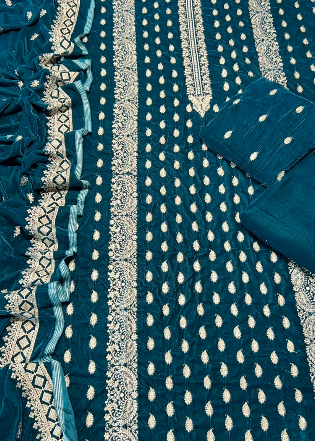 A close-up of a teal blue fabric textile with white embroidered patterns, borders, and matching folded fabric pieces arranged on the right side.