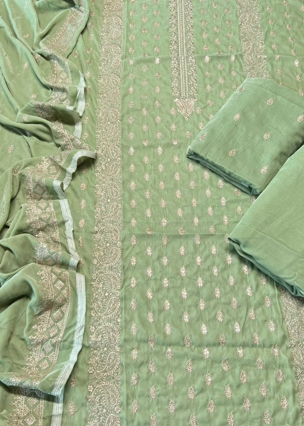 A sage green salwar suit set laid out flat, featuring a printed fabric body, matching pleated dupatta, and two folded fabric pieces with embroidered borders.
