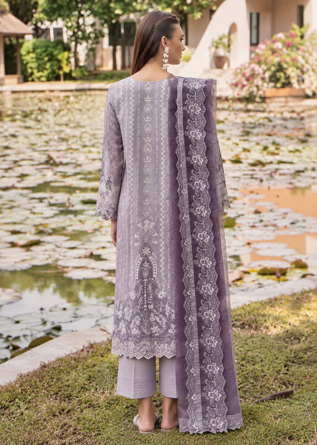 A woman stands outdoors by a pond wearing a lavender three-piece traditional outfit with embroidered kurta, matching pants, and a sheer embroidered dupatta.