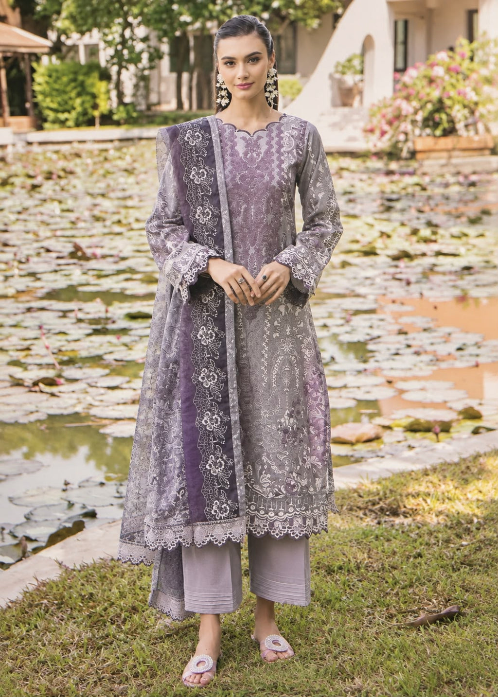 A woman stands outdoors in front of a pond with lily pads, wearing a lavender and gray embroidered traditional outfit with a matching dupatta and light gray pants.