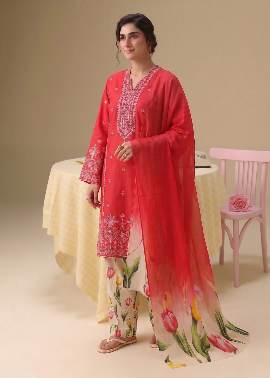A woman models a traditional three-piece salwar kameez suit in a studio setting. The outfit features a bright coral-red kameez jacket with white floral embroidery, matching printed trousers, and a sheer red dupatta with a tulip gradient print.