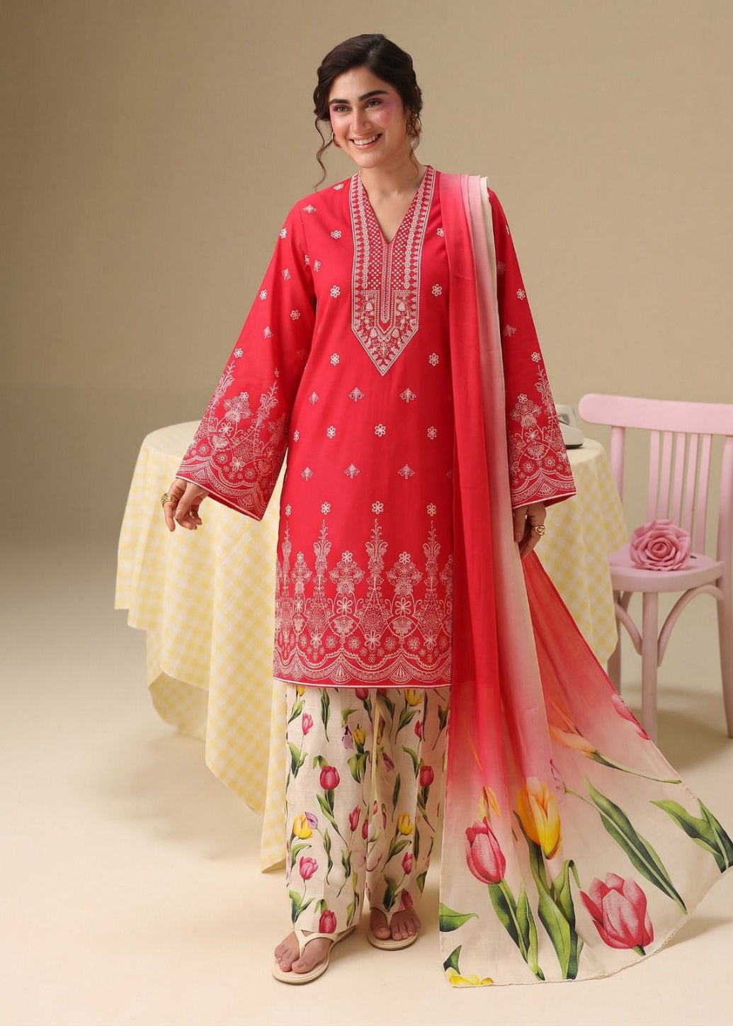 A woman modeling a traditional South Asian salwar kameez suit. The outfit features a bright coral-pink kurta with intricate white embroidery, matching floral-printed wide-leg pants, and a sheer pink dupatta with a green and pink tulip floral design.
