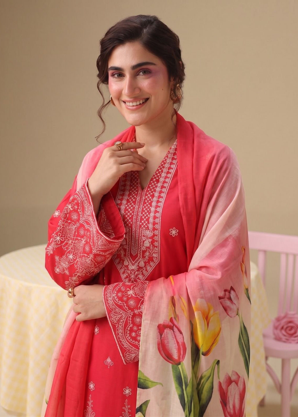 A woman wearing a traditional pink and red embroidered kurta with a floral printed dupatta, standing in a studio setting with a beige background and a pink chair.