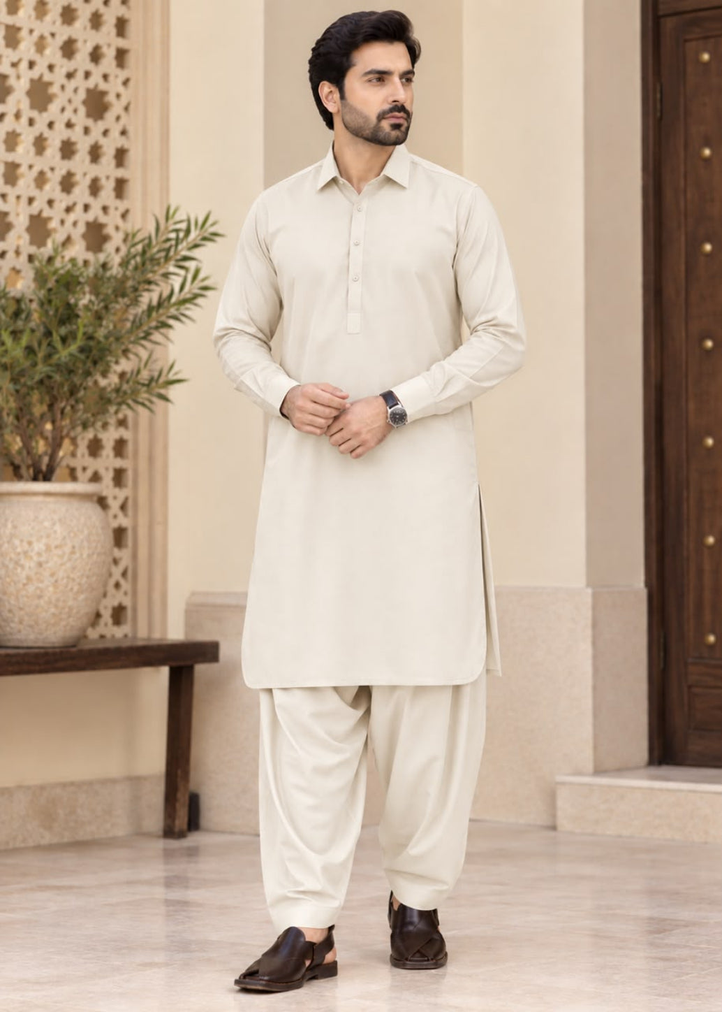 A man modeling a traditional light beige kurta and pant set in an elegant indoor setting with a patterned screen and potted plant.