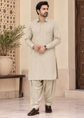 A man modeling a light beige long-sleeve kurta and matching trousers set, standing in a traditional architectural courtyard with a potted plant and wooden door in the background.