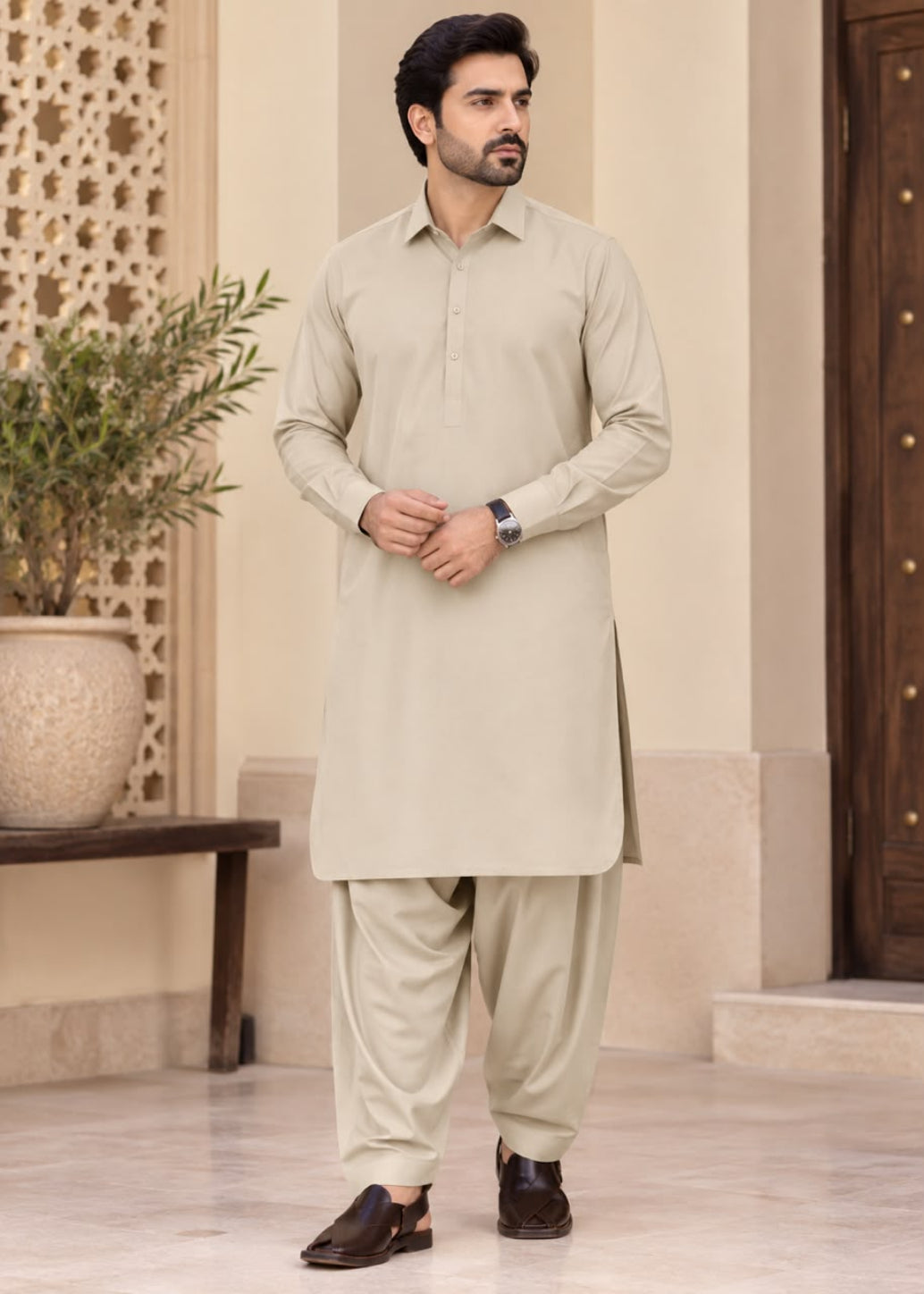 A man modeling a light beige long-sleeve kurta and matching trousers set, standing in a traditional architectural courtyard with a potted plant and wooden door in the background.