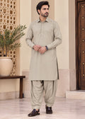 A man modeling a light beige traditional shalwar kameez suit with matching pants and dark brown leather loafers, standing in an indoor setting with a decorative screen and potted plant.