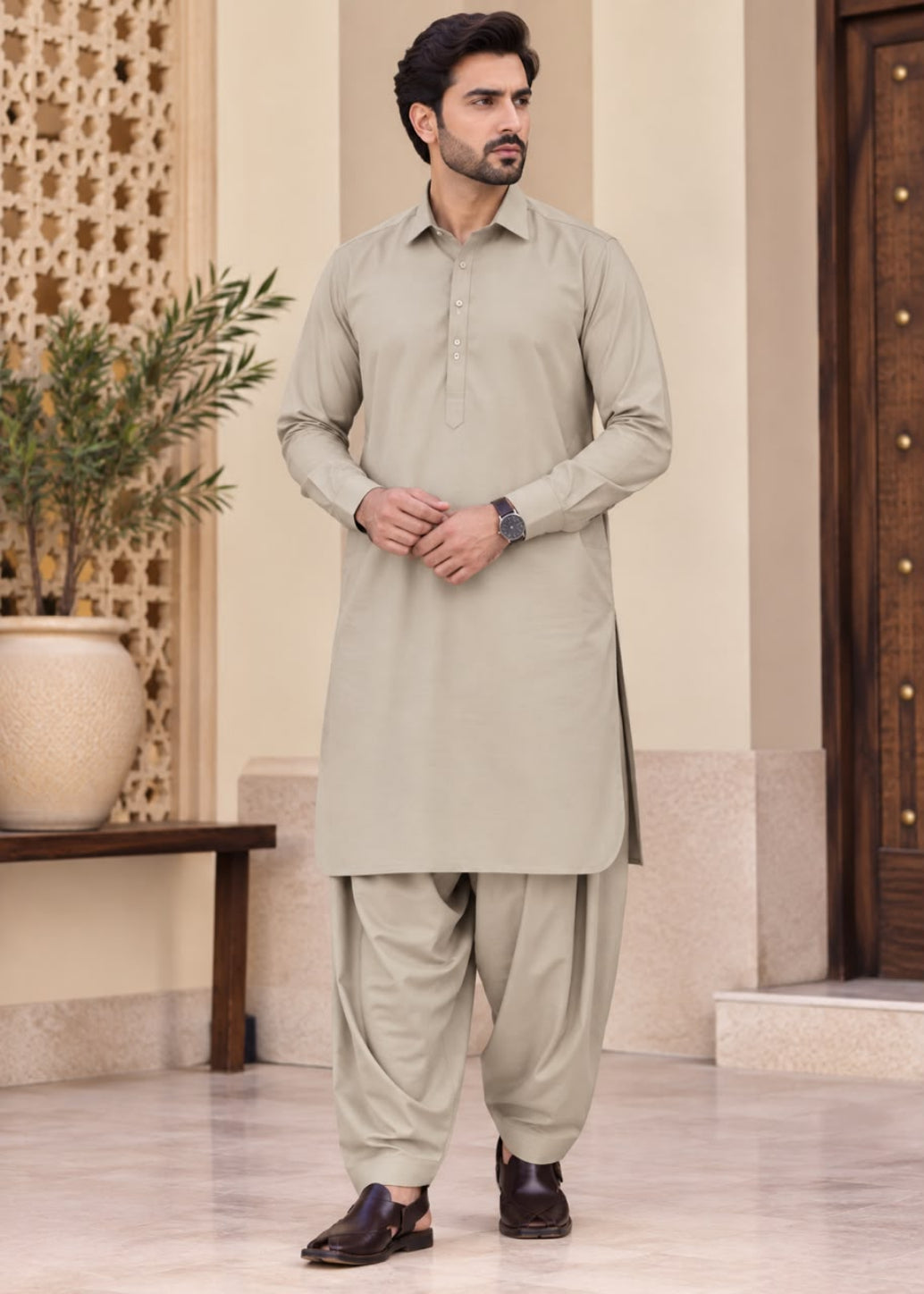 A man modeling a light beige traditional shalwar kameez suit with matching pants and dark brown leather loafers, standing in an indoor setting with a decorative screen and potted plant.