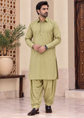 A man modeling a light olive green traditional South Asian outfit consisting of a long-sleeve kurta and matching loose trousers, standing in an indoor setting with a beige wall, patterned screen, and wooden door.