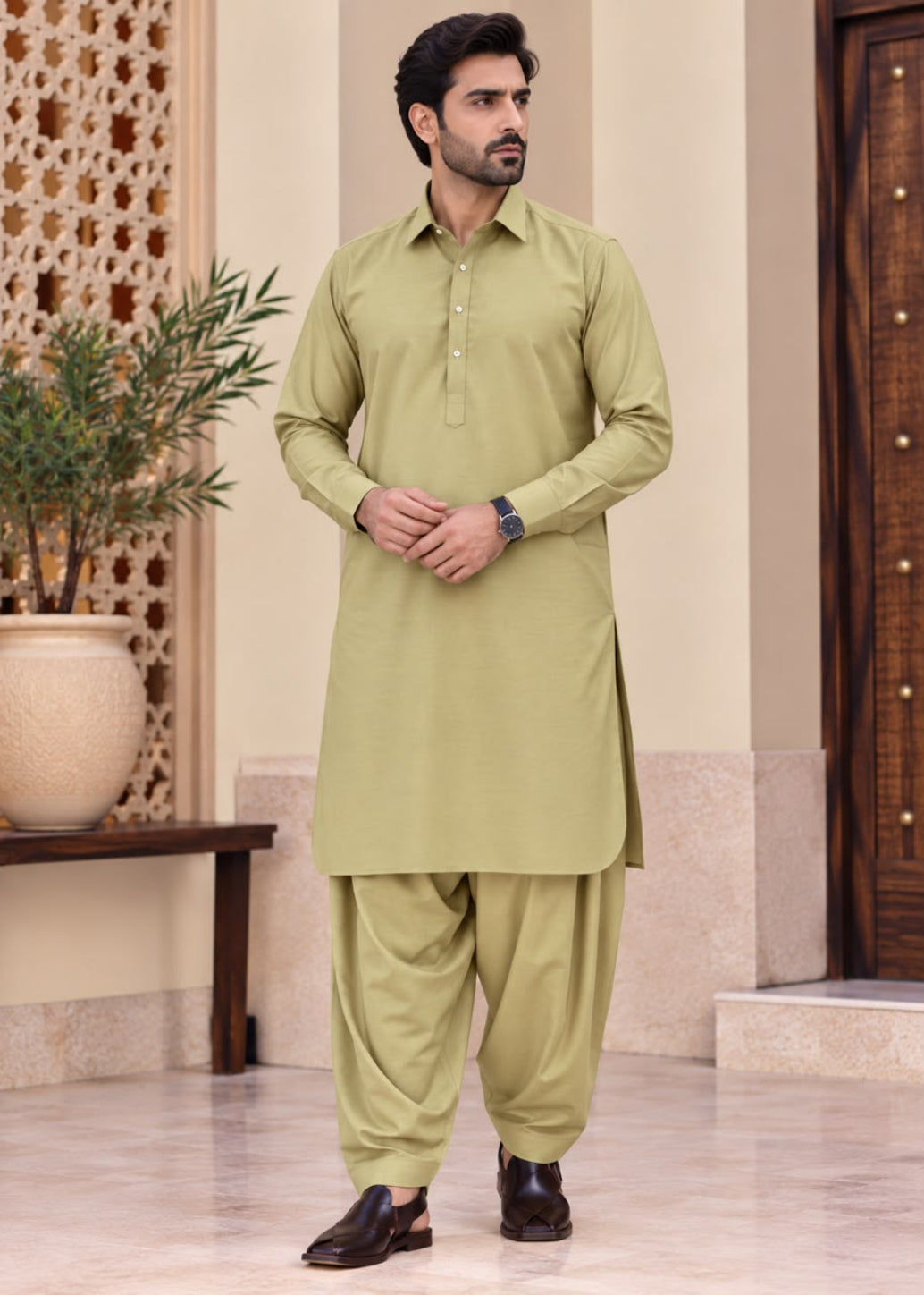 A man modeling a light olive green traditional South Asian outfit consisting of a long-sleeve kurta and matching loose trousers, standing in an indoor setting with a beige wall, patterned screen, and wooden door.