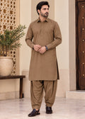 A man modeling a traditional brown three-piece shalwar kameez set, featuring a long-sleeve button-up kurta, matching pants, and brown leather shoes, standing in an elegant indoor setting.
