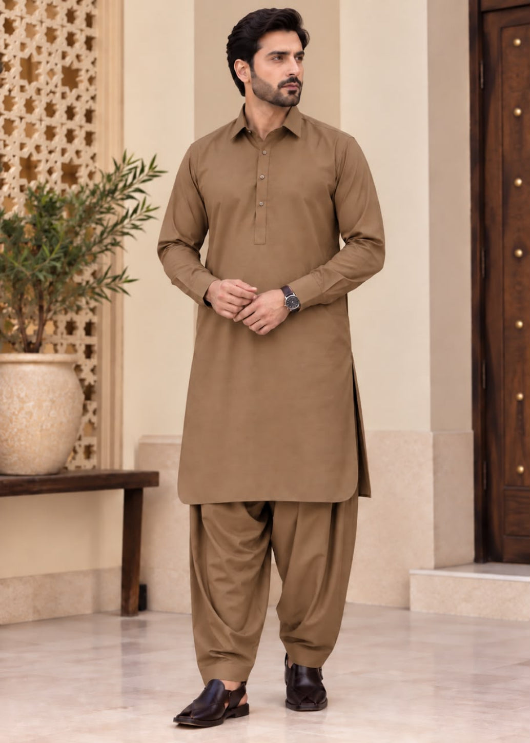 A man modeling a traditional brown three-piece shalwar kameez set, featuring a long-sleeve button-up kurta, matching pants, and brown leather shoes, standing in an elegant indoor setting.