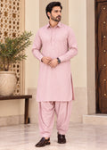 A man modeling a light pink traditional Indian outfit consisting of a long-sleeve kurta and matching pants, standing in an indoor setting with a beige wall, wooden door, and potted plant.