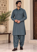 A man modeling a dark slate blue-grey traditional South Asian outfit consisting of a long-sleeve kurta and matching wide-leg trousers, standing in an indoor setting with a potted plant and wooden door in the background.