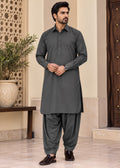 A man modeling a dark gray long-sleeve kurta and matching pants set, standing in a traditional indoor setting with beige walls and wooden door details.