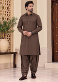 A man modeling a brown long-sleeve kurta and matching wide-leg pants set, standing in an indoor architectural setting with beige walls, a decorative screen, and a potted plant.