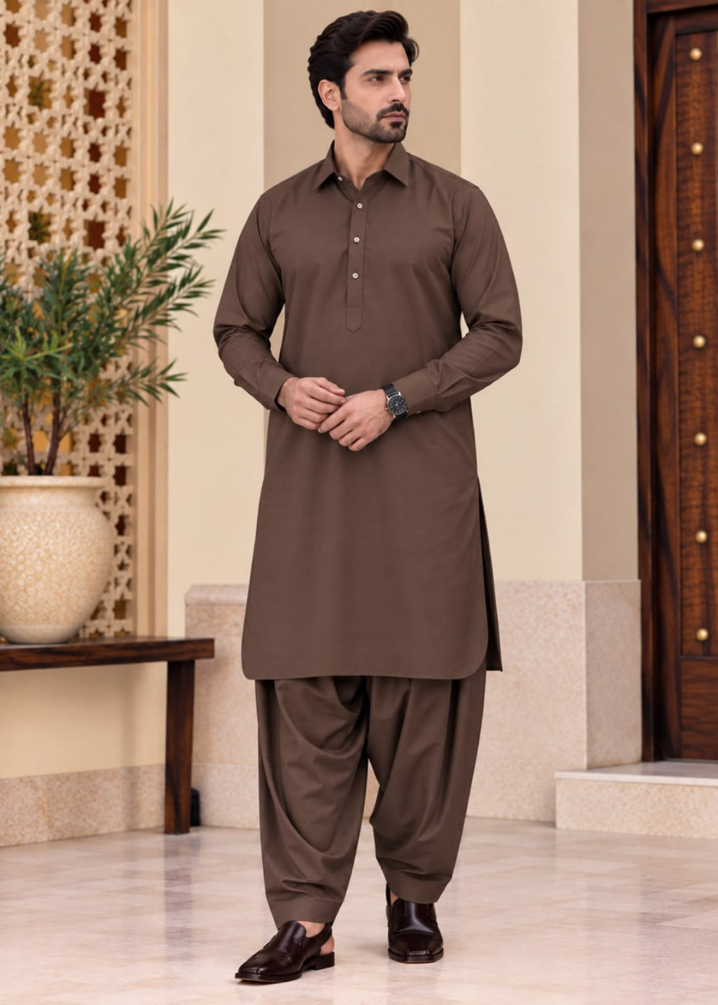 A man modeling a brown long-sleeve kurta and matching wide-leg pants set, standing in an indoor architectural setting with beige walls, a decorative screen, and a potted plant.