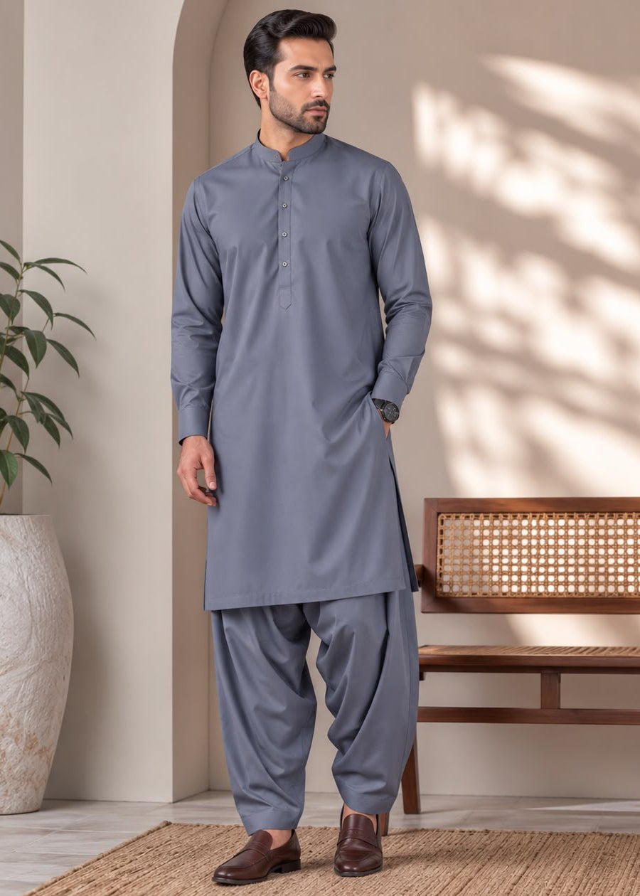 A man modeling a traditional Indian outfit consisting of a gray long-sleeve kurta and matching pants, standing in a styled indoor setting with a bench, potted plant, and natural light shadows.