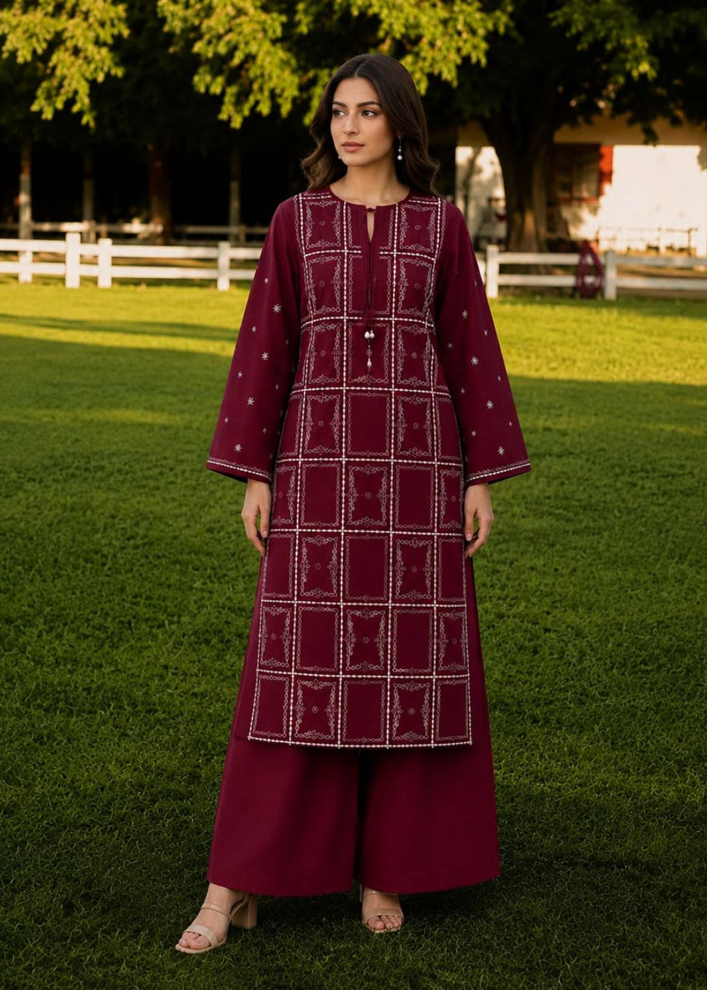 A woman stands on a green lawn wearing a deep maroon embroidered kurta with matching wide-leg trousers. The kurta features white geometric patterns and a central embroidered panel, paired with a matching maroon tunic underneath.