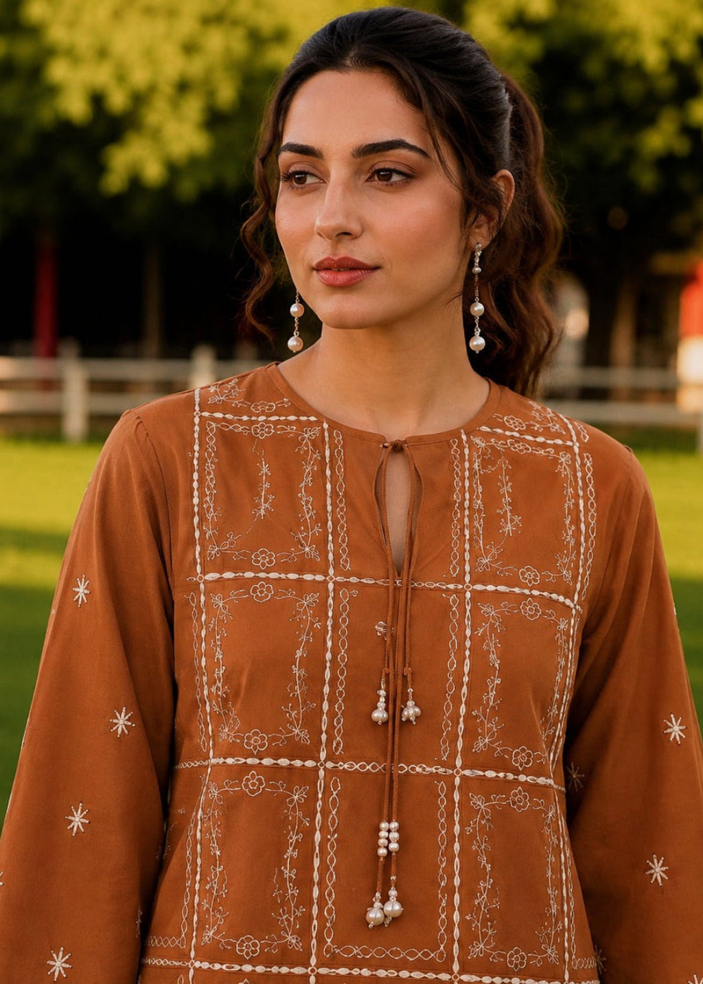 A woman wearing a rust-brown embroidered kurta with white geometric and floral patterns, standing outdoors in a grassy park setting.