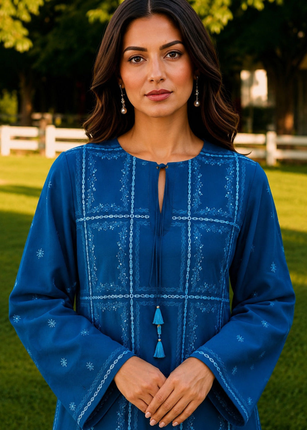 A woman wearing a vibrant blue long-sleeve embroidered tunic with white geometric patterns and tassel details, standing in a sunny grassy park.