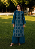 A woman stands on a grassy lawn wearing a teal blue embroidered kurta and matching wide-leg pants. The outfit features white geometric patterns and small embroidered details.