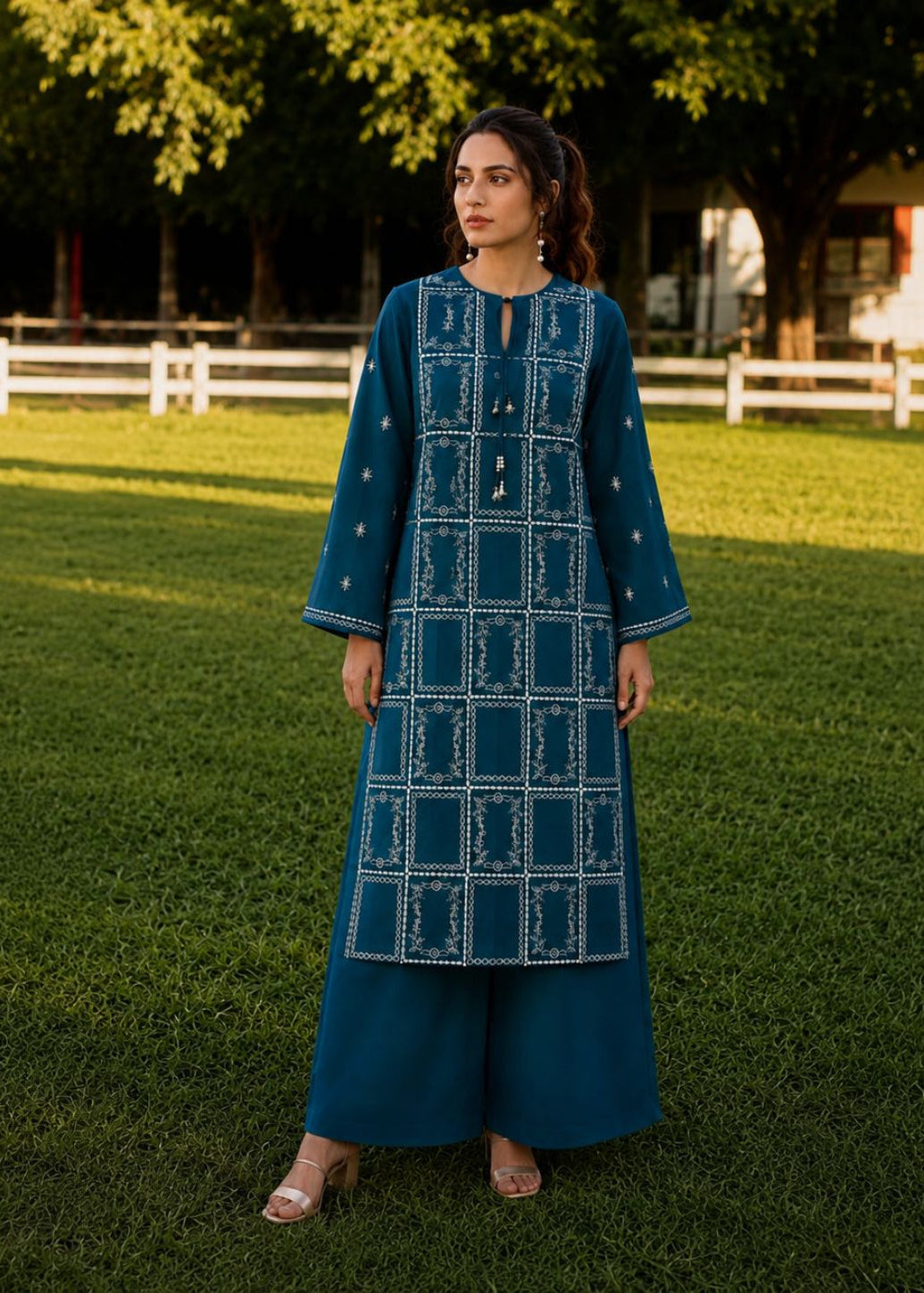 A woman stands on a grassy lawn wearing a teal blue embroidered kurta and matching wide-leg pants. The outfit features white geometric patterns and small embroidered details.