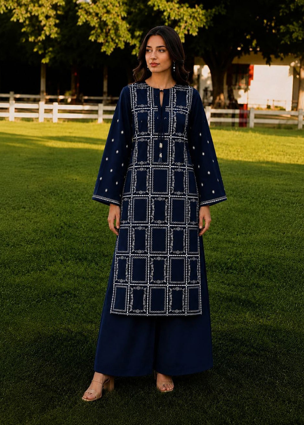 A woman stands on a green lawn wearing a navy blue embroidered kurta with wide sleeves and matching wide-leg pants. The outfit features white geometric embroidery and small white dot details.