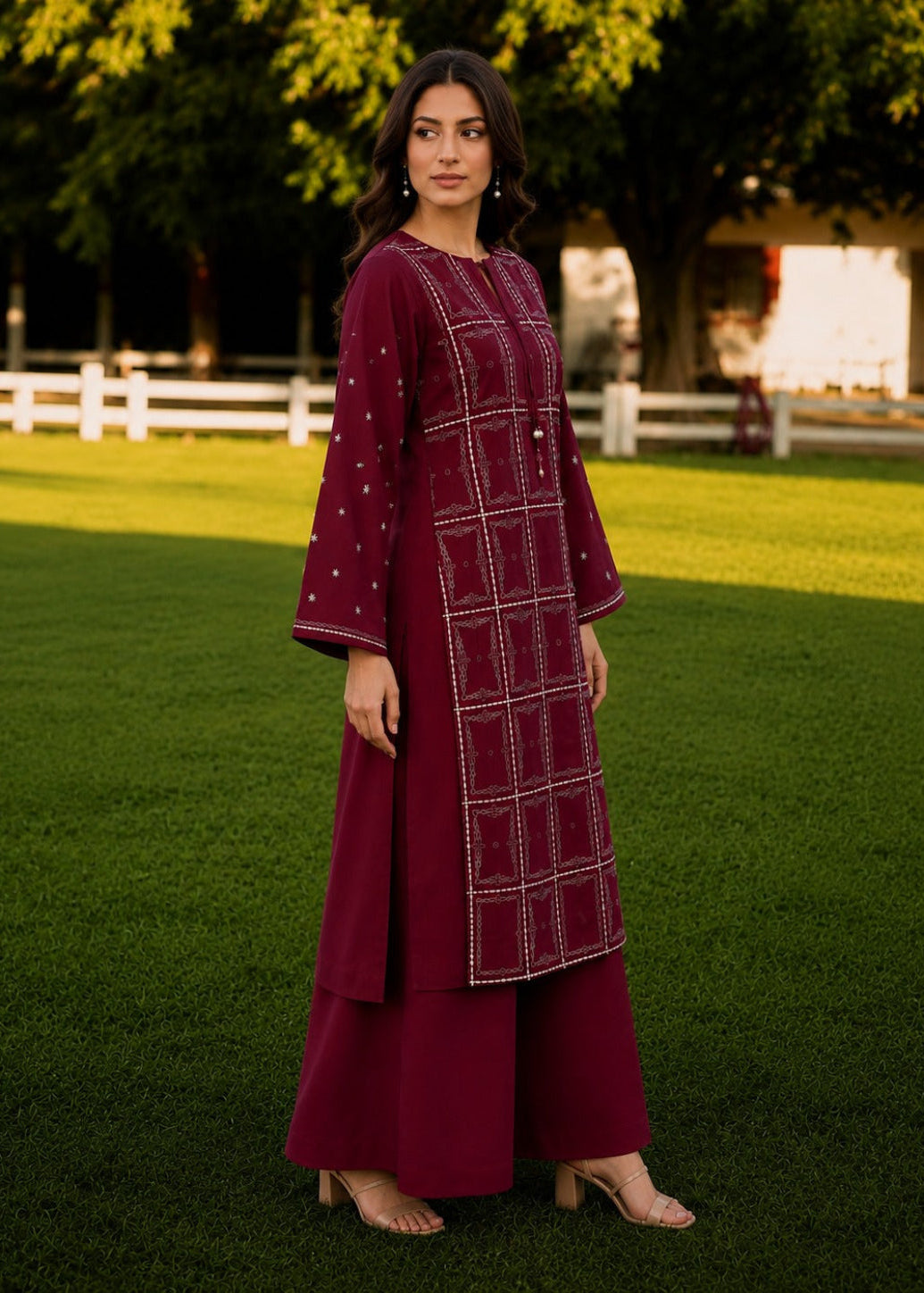 A woman stands on a green lawn wearing a deep maroon traditional outfit with white embroidered geometric patterns and matching wide-leg pants. Trees and a white fence are visible in the background.