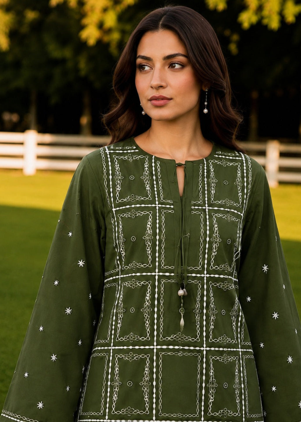 A woman wearing an olive green embroidered tunic with white geometric patterns and star details, standing outdoors in a grassy park setting.