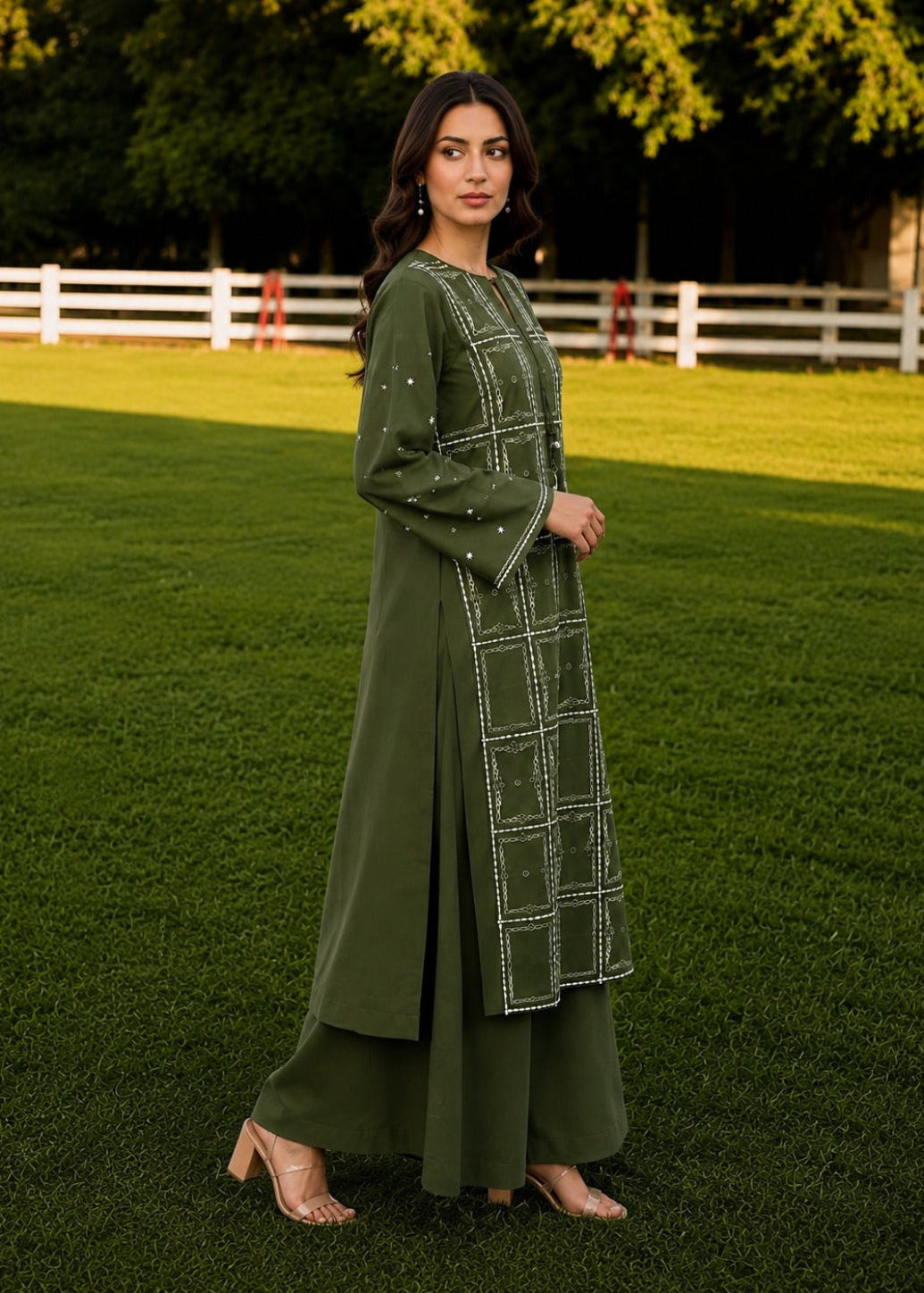 A woman stands on a green lawn wearing an olive green long kurta with white geometric embroidery and matching wide-leg pants. The background features a white fence and trees.