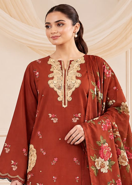 A woman models a rust-red ethnic three-piece suit set featuring a kurta with gold embroidery, matching pants, and a floral printed dupatta, set against a cream-colored paneled wall.