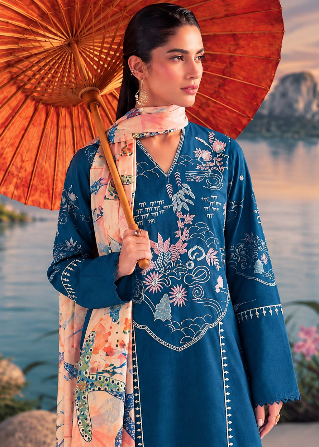 A woman models a traditional blue embroidered kurta with a matching printed dupatta, holding a large orange paper umbrella against a scenic water backdrop.