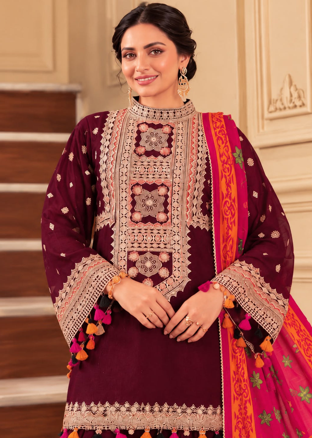 A woman models a traditional Indian salwar kameez suit in deep maroon with intricate gold and silver embroidery, paired with a bright pink and orange patterned dupatta, set against an elegant indoor background with beige walls and wooden stairs.