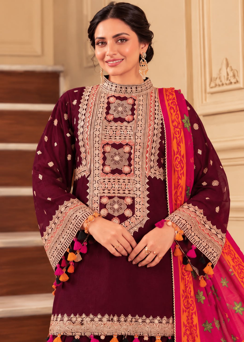A woman models a traditional Indian salwar kameez suit in deep maroon with intricate gold and silver embroidery, paired with a bright pink and orange patterned dupatta, set against an elegant indoor background with beige walls and wooden stairs.