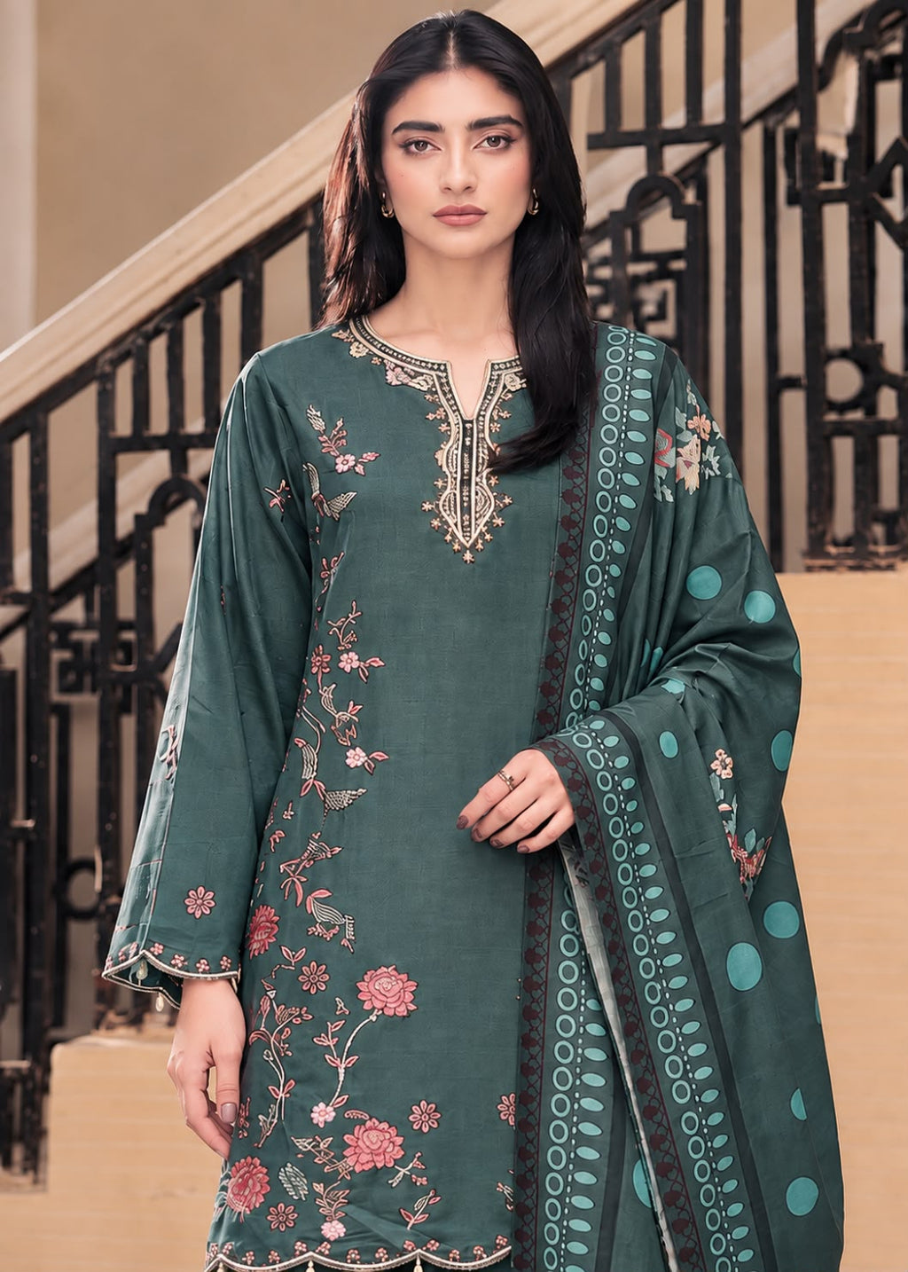 A woman models a dark teal green 3-piece salwar kameez suit with floral embroidery and a matching dupatta in an elegant indoor setting with a staircase.
