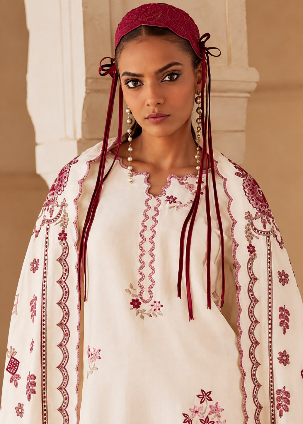 A woman wearing a cream-colored embroidered traditional outfit with deep red embroidery, a matching red headscarf, and long dangling earrings, posed against a warm beige architectural background.