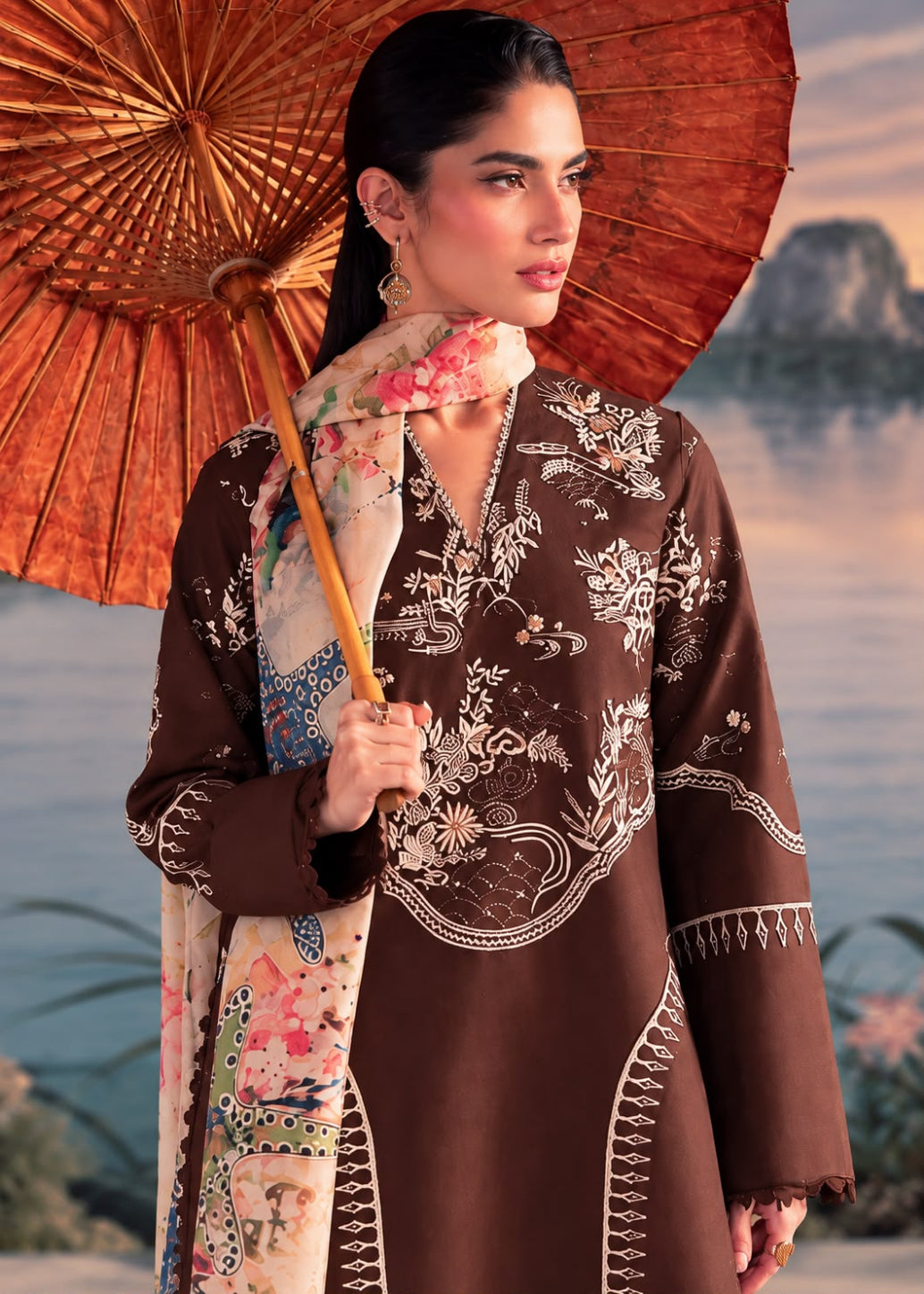 A woman models a brown embroidered kurta with a floral printed dupatta, holding a traditional brown paper umbrella against a scenic water backdrop.