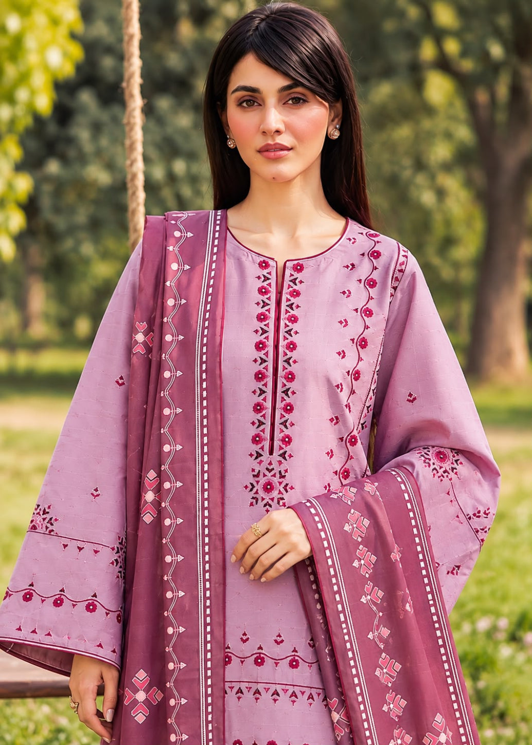 A woman wearing a lavender purple embroidered traditional outfit with a matching dupatta, standing outdoors in a park setting.
