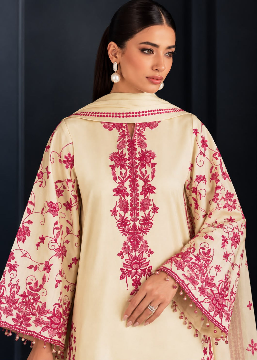 A woman modeling a cream-colored traditional outfit with vibrant magenta embroidery on the sleeves, collar, and back, set against a dark studio background.